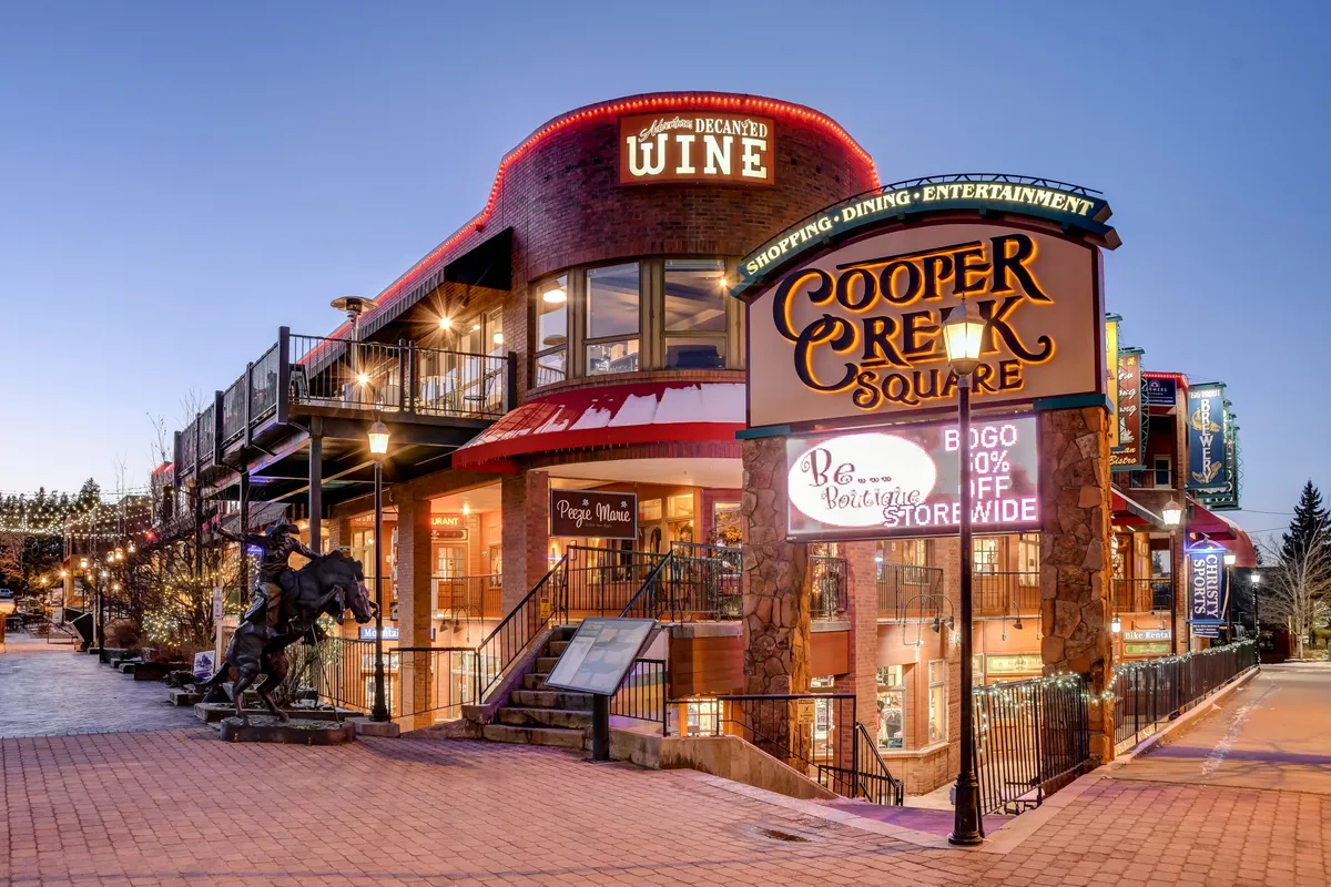 A festive downtown scene at Cooper Creek Square with visible outdoor dining areas, boutique stores, community signage, and a prominent horse statue. The space buzzes with the possibility of shopping, dining, and local events, set against a backdrop of mountain-town charm.