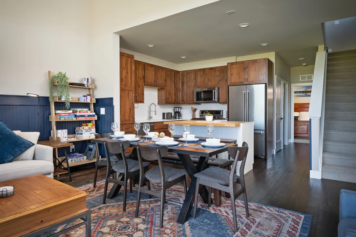 A warm, inviting dining area with a table set for a meal, surrounded by comfortable seating. The space flows seamlessly into a well-appointed kitchen with rich wooden cabinetry and a nearby living area, illustrating the open-concept design. The setup invites communal dining and casual conversation.