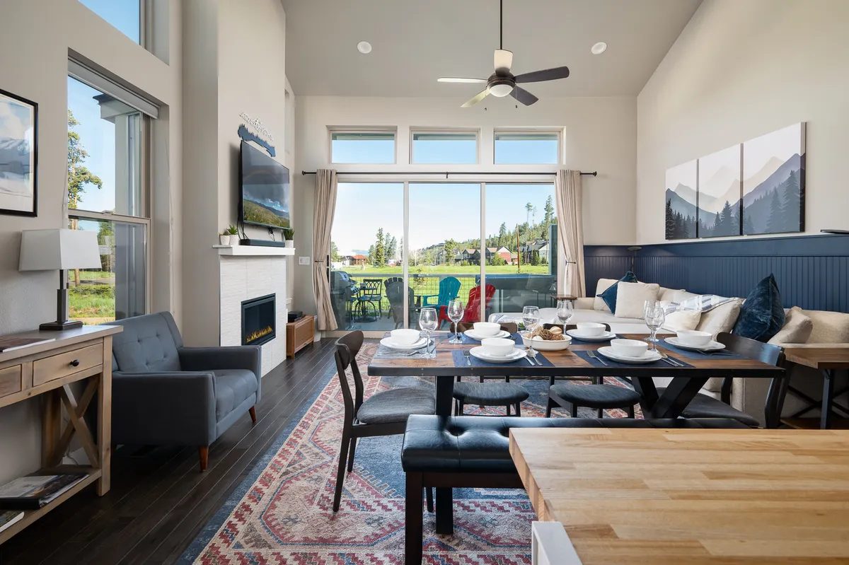 A bright, open dining and living area features a welcoming dining table set with dishes, comfortable seating, and large windows that offer scenic mountain views. The space exudes warmth and invites family gatherings, meals, and relaxation by the fireplace—all in a setting that merges comfort and elevated style.
