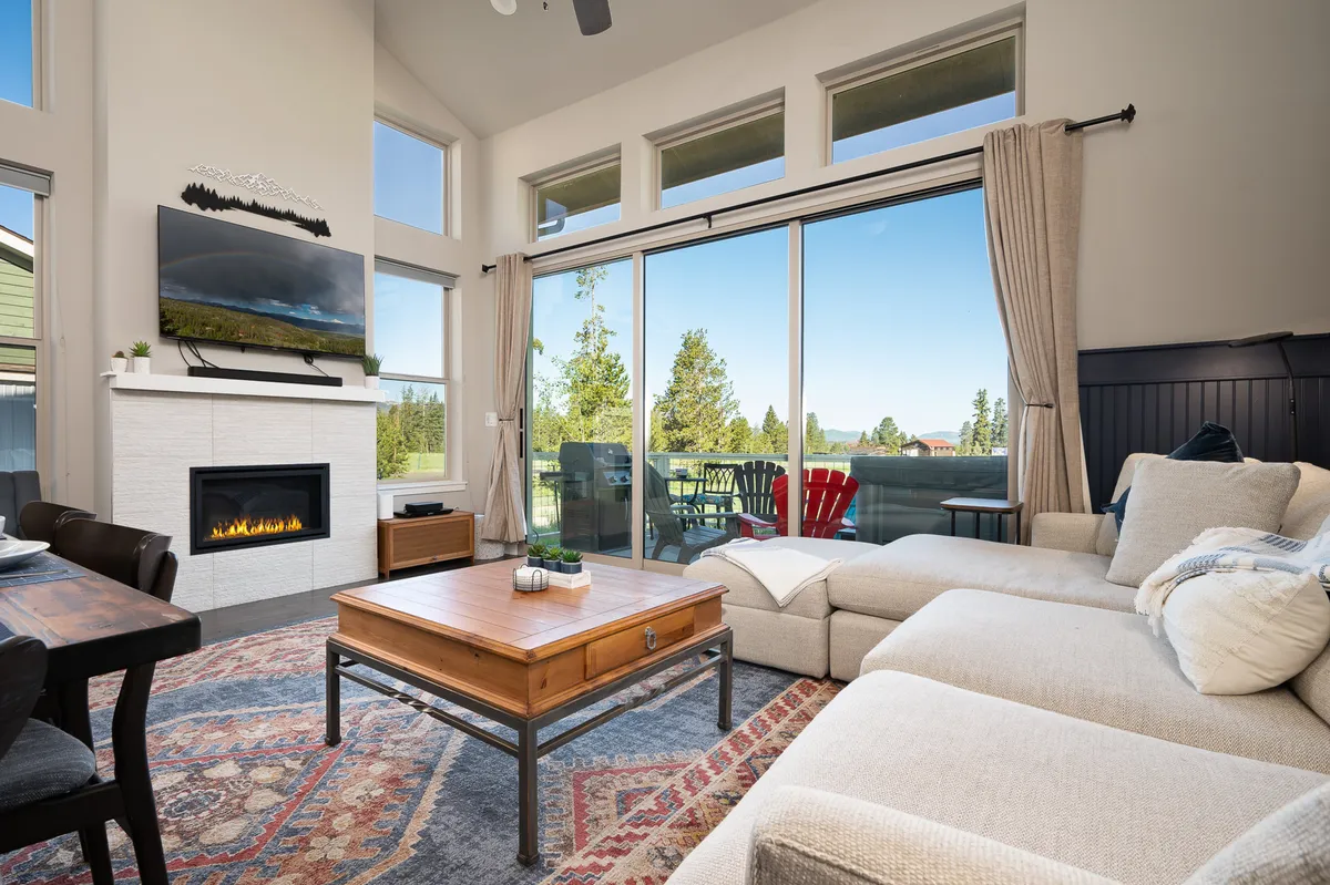 A cozy living room with a plush sofa and armchairs centered around a modern gas fireplace, large windows letting in abundant natural light, and a coffee table atop a textured rug. The mounted TV is visible above the fireplace, and the space feels both intimate and open, perfect for gathering and relaxing after outdoor adventures.