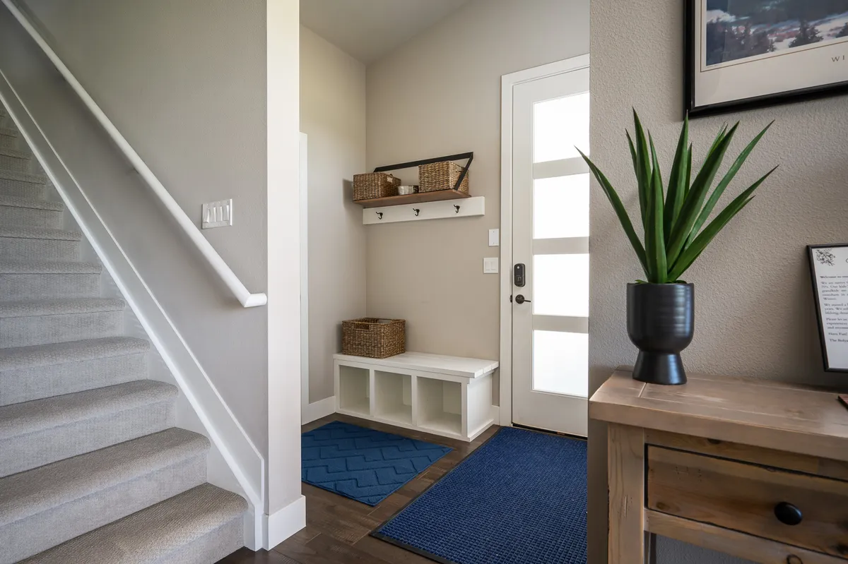 A welcoming entryway featuring a door with glass panels, an entry bench, blue rugs, and an indoor plant. The space radiates warmth and invites guests into the home, suggesting both comfort and an inviting atmosphere.