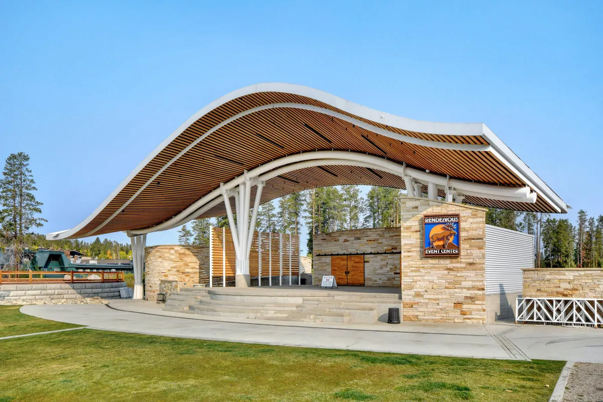 This image shows the distinctive curved wooden canopy and stone facade of Hideaway Park, complete with event signage. The setting is outdoors, with clear elements indicating it's a venue for community events such as concerts, festivals, and gatherings.