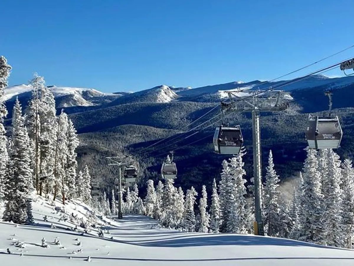 A panoramic view of ski gondolas gliding above snow-covered slopes, backed by a striking Rocky Mountain range and bordered by evergreen trees. The scene exudes the allure of alpine adventure and tranquility.