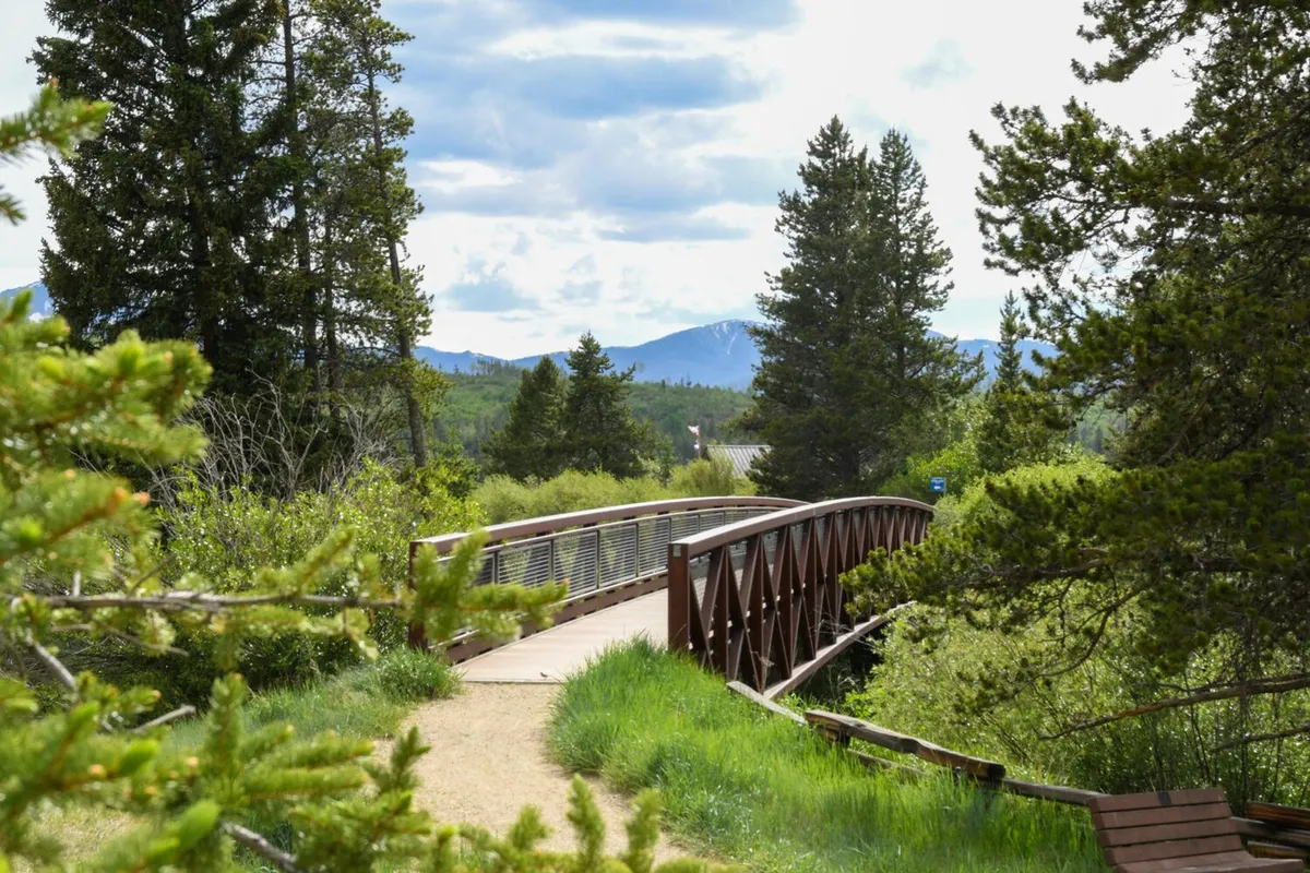 An inviting wooden bridge extends over a lush natural landscape, framed by dense green trees and distant mountains. The bridge is surrounded by natural trails, perfect for walking or biking, and the scenic mountain backdrop evokes the spirit of outdoor adventure.