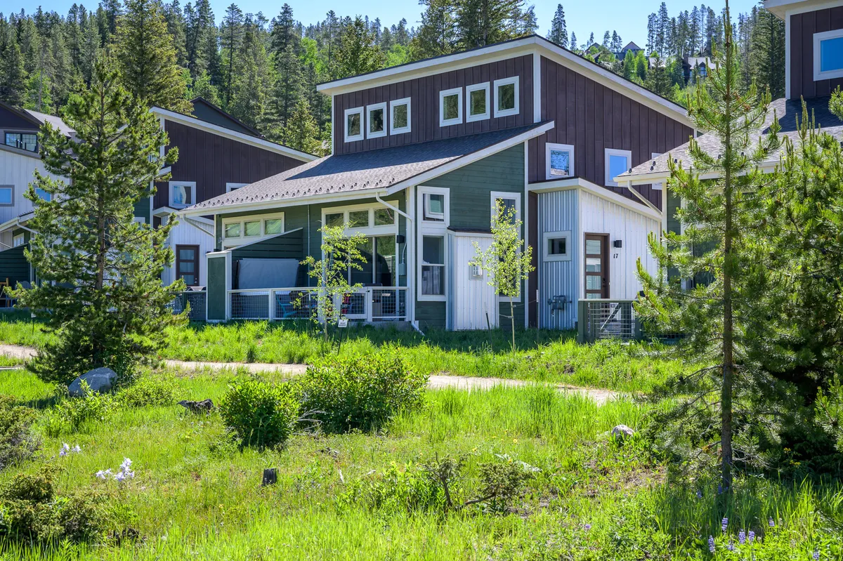 The hero image showcases a striking modern mountain cabin nestled amidst lush greenery in Fraser, Colorado. The home's sleek architectural lines are complemented by a welcoming pathway leading to the entrance, with natural landscaping surrounding the property. The inviting porch offers a perfect vantage point to take in mountain sunsets, while the serene outdoor setting hints at the possibility of nature walks and wildlife exploration right outside the door.
