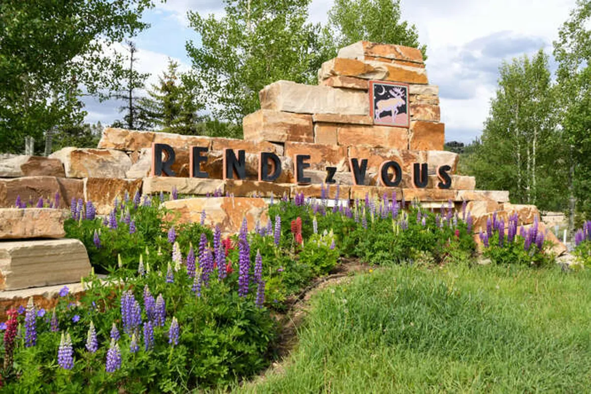 A stone monument marked 'RENDEZVOUS' is surrounded by vibrant flower beds and mature trees, suggesting a well-maintained, welcoming community space. The landscaping provides an inviting area for both humans and pets to explore and enjoy the outdoors.